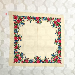 Op de afbeelding: Cr&egrave;mekleurig vierkant textiel met een bloemenrand. De rand heeft rode rozen, blauwe bloemen en groene bladeren. Het textiel is waarschijnlijk een sjaal of een decoratief stuk.