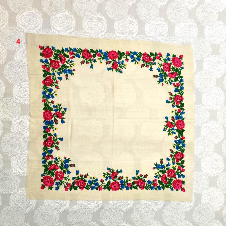 Op de afbeelding: Cr&egrave;mekleurig vierkant textiel met een bloemenrand. De rand heeft rode rozen, blauwe bloemen en groene bladeren. Het textiel is waarschijnlijk een sjaal of een decoratief stuk.