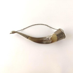 Op de afbeelding: Een gebogen hoornen vat met een gepolijst, meerkleurig bruin en crèmekleurig oppervlak. Het object is voorzien van een zilverkleurige metalen rand en een punt, verbonden door een dunne, delicate zilverkleurige metalen ketting om op te hangen.