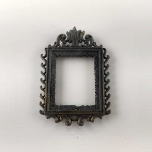 May include: Ornate, dark bronze-colored picture frame with a rectangular opening. The frame features detailed scrollwork and a decorative top element. The frame is likely made of metal and is designed for a small photograph or artwork.