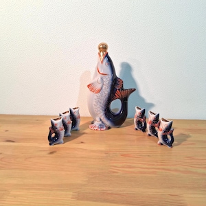 Puede incluir: Un juego de licorera de cerámica con forma de pez con seis vasos de chupito a juego. La licorera es blanca y azul con detalles en naranja y un tapón dorado. Los vasos también tienen forma de pez.