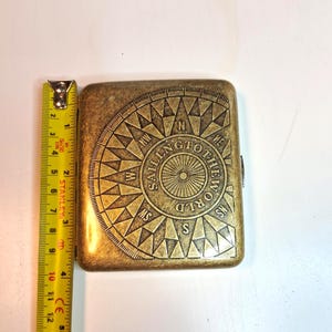 K&ouml;nnte beinhalten: Ein rechteckiges, antik-messingfarbenes Etui mit einem Kompassrosen-Design und dem Schriftzug "SAILING TO THE WORLD". Das Etui ist etwa 10 cm lang. Die Kompassrose hat Richtungsanzeigen.