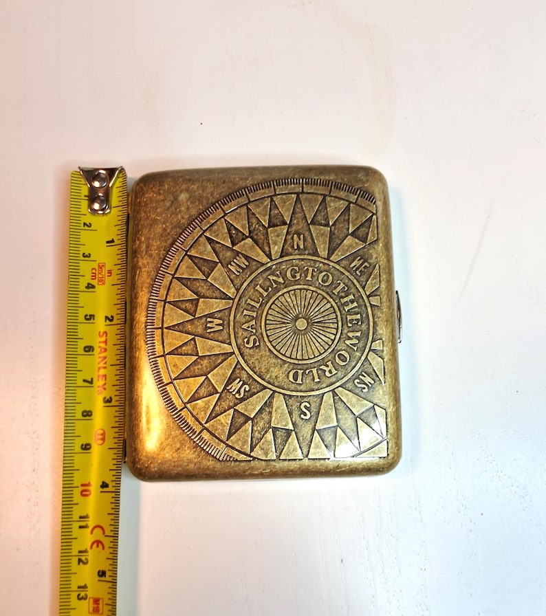 K&ouml;nnte beinhalten: Ein rechteckiges, antik-messingfarbenes Etui mit einem Kompassrosen-Design und dem Schriftzug "SAILING TO THE WORLD". Das Etui ist etwa 10 cm lang. Die Kompassrose hat Richtungsanzeigen.
