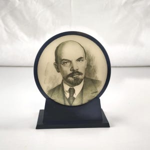 May include: A circular desk ornament featuring a black and white portrait of Vladimir Lenin. The portrait is encased in a dark blue circular frame that rests on a matching dark blue rectangular base, creating a stable display piece for a desk.