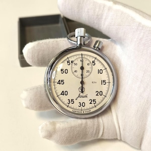 May include: A vintage chrome stopwatch with a white face, black numerals, and a small star emblem. The stopwatch is held in a gloved hand, with a gray case visible in the background. The text "СДЕЛАНО В СССР" is printed on the face.
