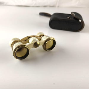 May include: Ivory and gold opera glasses with a black leather case. The vintage binoculars have a cream-colored body with gold-toned accents around the lenses. The case is rectangular with a snap closure and a small handle.