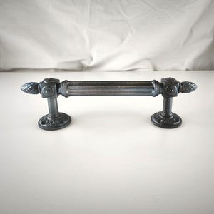 Könnte beinhalten: Ein dunkelgrauer Möbelgriff aus Metall mit einer zentralen zylindrischen Stange mit vertikalen Rillen, flankiert von quadratischen Blöcken mit Blumenmotiven und spitzen Endstücken, getragen von zwei runden Montagebasen.