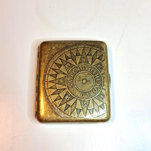 K&ouml;nnte beinhalten: Ein rechteckiges Zigarettenetui in antikem Messingfarbton. Das Etui zeigt ein Kompassrosen-Design mit den Worten "SAILING TO THE WORLD" in einem kreisf&ouml;rmigen Muster. Das Etui ist geschlossen und hat einen Klappverschluss.