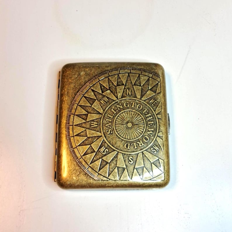 K&ouml;nnte beinhalten: Ein rechteckiges Zigarettenetui in antikem Messingfarbton. Das Etui zeigt ein Kompassrosen-Design mit den Worten "SAILING TO THE WORLD" in einem kreisf&ouml;rmigen Muster. Das Etui ist geschlossen und hat einen Klappverschluss.