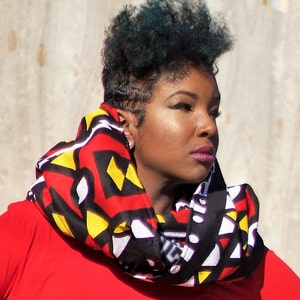 May include: A colorful infinity scarf with a geometric pattern in red, black, yellow, and white. The scarf is wrapped around the neck, creating a bold and stylish accessory. The scarf is made of a soft material.