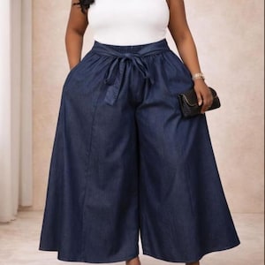 May include: A white tank top paired with wide-leg, dark blue denim pants. The pants feature a tie-waist detail and pockets. The outfit is completed with black sandals and a small black clutch.
