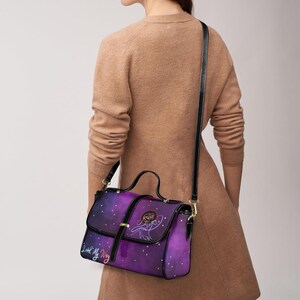 Peut inclure: Sac &agrave; main violet et bleu avec un motif galaxie et une bandouli&egrave;re noire. Le sac est dot&eacute; d'une fermeture &agrave; rabat, d'une poign&eacute;e sup&eacute;rieure et d'une bandouli&egrave;re. Le sac porte un dessin d'une personne et le texte "Look My Way".