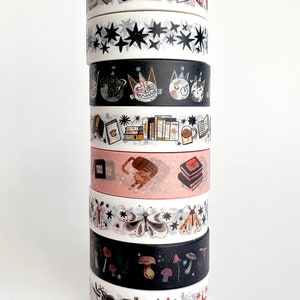 May include: A stack of eight washi tapes with various designs, including stars, cats, books, butterflies, mushrooms, and a forest scene. The tapes are all white with black and colored designs.