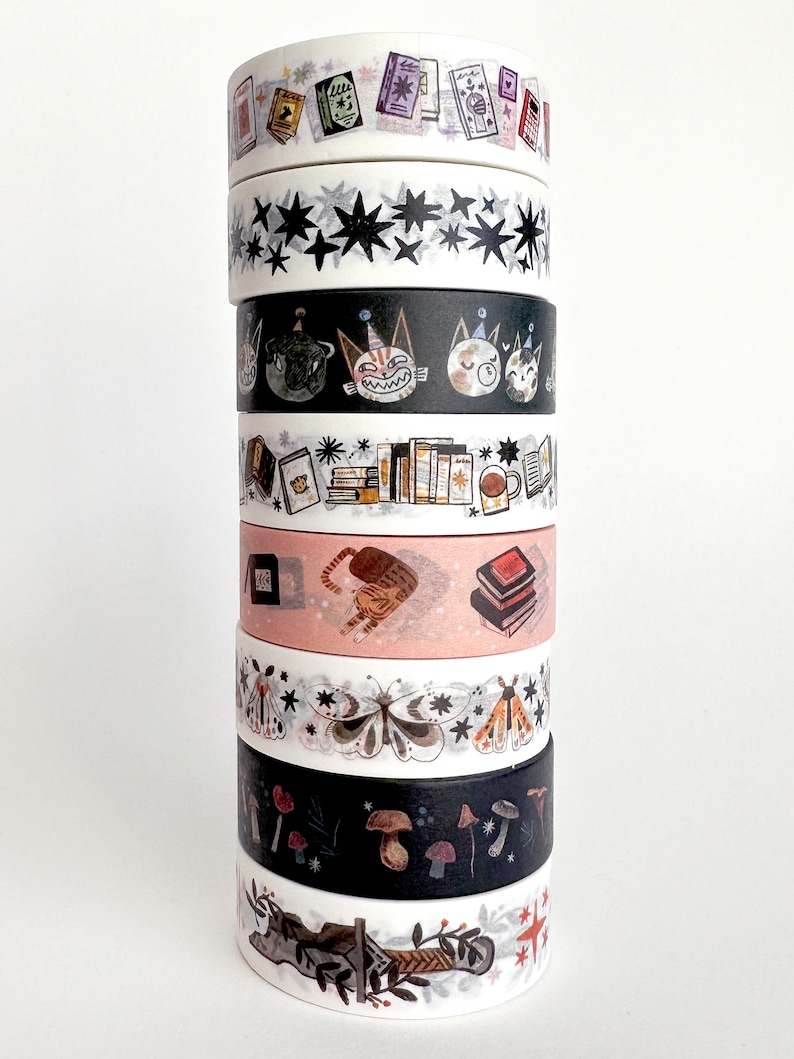 May include: A stack of eight washi tapes with various designs, including stars, cats, books, butterflies, mushrooms, and a forest scene. The tapes are all white with black and colored designs.