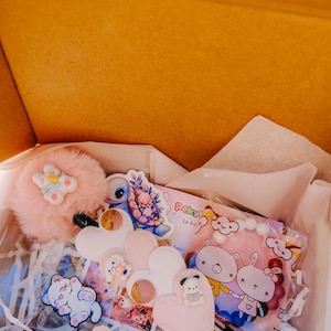 Peut inclure: Une boîte en carton ouverte remplie de divers accessoires mignons. On y trouve un pompon rose duveteux avec un ours, des autocollants, un support de téléphone en forme de cœur et une pochette rose avec des ours et un lapin de dessin animé.