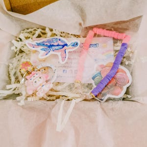 Può includere: Una scatola contenente vari oggetti a tema Kawaii. Include un adesivo a forma di tartaruga blu e rosa, braccialetti rosa e viola e un ciondolo rosa e oro. Gli articoli sono confezionati con carta velina e carta sminuzzata.