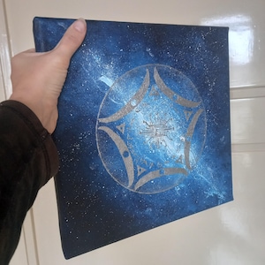Op de afbeelding: Vierkante canvas kunst met een donkerblauw en wit melkwegstelsel ontwerp. Een zilveren geometrische vorm met een printplaatontwerp staat in het midden. Het kunstwerk wordt door een persoon vastgehouden.