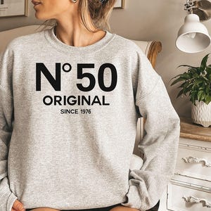 May include: A light gray crewneck sweatshirt featuring black screen-printed text on the chest. The design displays the characters N°50 in a large font, with the word ORIGINAL below it and the text SINCE 1976 at the bottom.