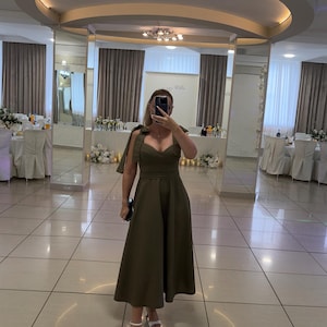 May include: An olive green midi dress with a sweetheart neckline and shoulder tie details. The dress has a fitted bodice and a flared skirt. The person is wearing white heeled sandals. The background is a well-lit room with mirrors and tables.