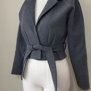 May include: A gray wrap jacket with a tie belt, displayed on a mannequin. The jacket features a notched collar and long sleeves. The tie belt cinches at the waist, creating a flattering silhouette. A stylish outerwear piece.