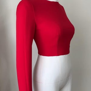 May include: A bright red long-sleeved crop top on a white dress form. The top has a crew neck and a fitted shape, demonstrating a tailored design. The dress form is set against a plain white background.
