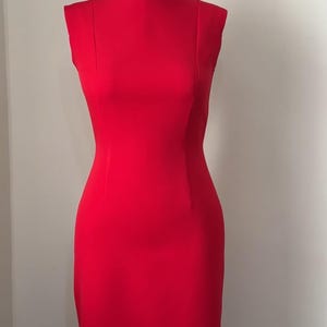 Könnte beinhalten: Ein ärmelloses, knielanges rotes Kleid, das auf einer Schaufensterpuppe präsentiert wird. Das Kleid hat eine figurbetonte Silhouette mit rundem Ausschnitt und dezenten Nahtdetails. Die leuchtend rote Farbe und das klassische Design deuten auf formelle oder Business-Kleidung hin.