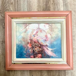 Könnte beinhalten: Gerahmtes Kunstwerk mit einem bunten Vogel in der Nähe eines dekorativen Vogelkäfigs. Das Gemälde hat eine weiche, verträumte Qualität mit Pastellfarben, darunter Rosa, Blau und Gold. Der Rahmen ist ein gedämpftes Rosa mit einem goldenen Akzent.