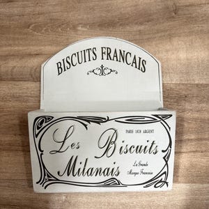 May include: White wooden wall-mounted storage box with the text "BISCUITS FRANCAIS" at the top and "Les Biscuits Milanais" below. The box has a decorative black border and text.