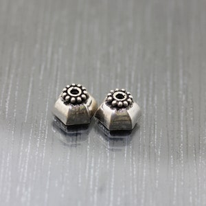 K&ouml;nnte beinhalten: Zwei silberfarbene Metallperlen mit einem dekorativen Design. Die Perlen haben eine runde Mitte mit einem Kreis aus kleinen Punkten um den Rand. Die Perlen sind sechseckig.