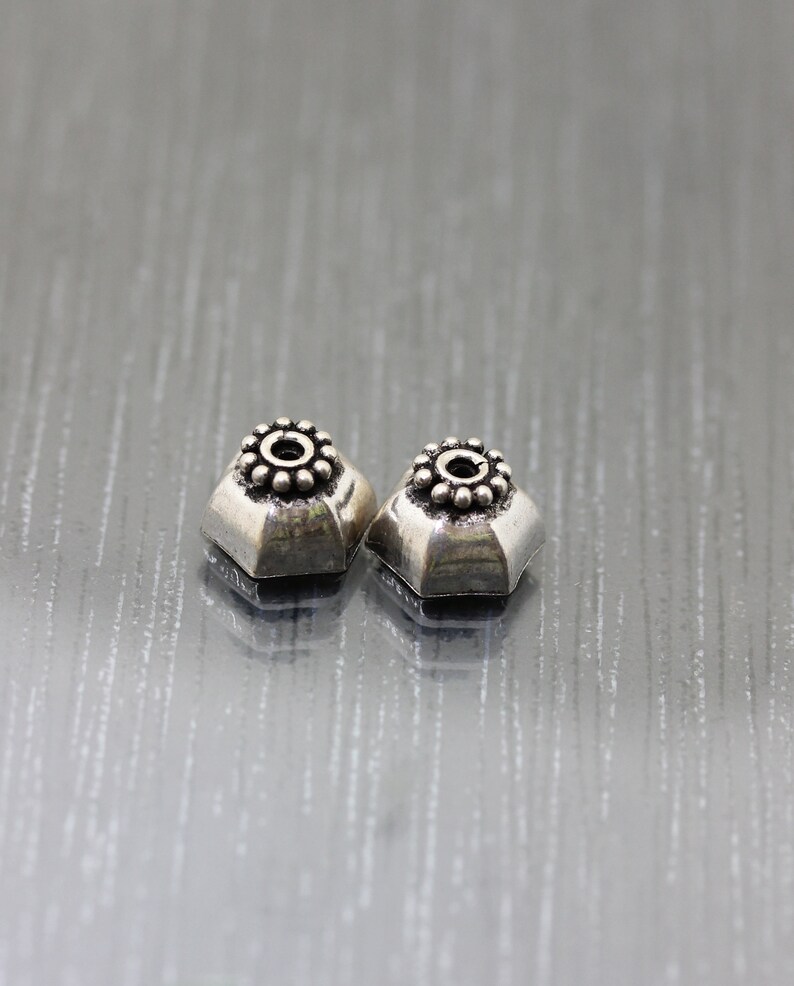 K&ouml;nnte beinhalten: Zwei silberfarbene Metallperlen mit einem dekorativen Design. Die Perlen haben eine runde Mitte mit einem Kreis aus kleinen Punkten um den Rand. Die Perlen sind sechseckig.