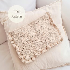 Könnte beinhalten: Eine cremefarbene Häkeltasche mit floralem Muster und Rüschenkanten liegt auf einem beigen Kissen. Die rechteckige Tasche hat ein detailliertes, offenes Muster. Der Text "PDF Pattern" befindet sich in einem Kreis oben links.