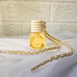 Puede incluir: Pequeña botella de vidrio facetado llena de líquido amarillo, con tapa de madera. Una cuerda trenzada dorada y blanca está unida. Probablemente un ambientador o perfume para colgar.