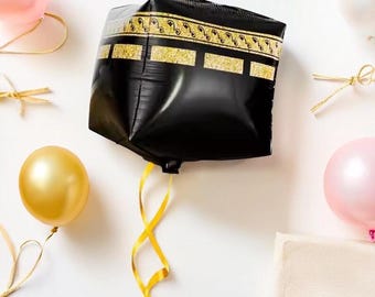 Puede incluir: Un globo de aluminio negro de forma cuadrada con una banda de purpurina dorada con un patrón repetitivo en la parte superior, suspendido por una cinta amarilla larga, rodeado de globos redondos más pequeños en rosa, dorado y blanco sobre un fondo liso.