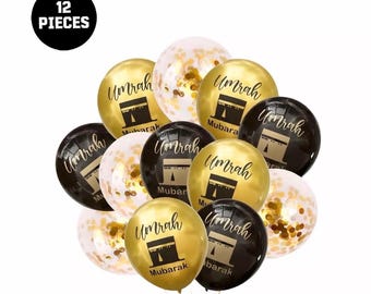 Puede incluir: Un conjunto de 12 globos dorados, negros y transparentes. Los globos dorados y negros tienen la palabra "Umrah" y un gráfico de un edificio. Algunos globos están llenos de confeti dorado.