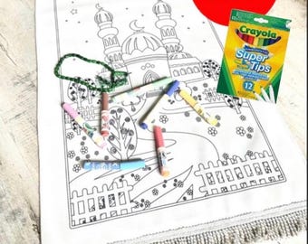 Puede incluir: Una alfombra de oración blanca para colorear con un diseño de mezquita, flecos y un juego de 12 rotuladores lavables Crayola Super Tips. La alfombra presenta un contorno negro de una mezquita, nubes y elementos florales.