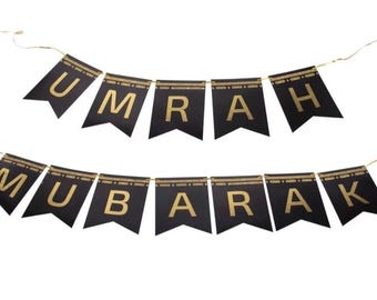 Puede incluir: Bandera negra con letras doradas que deletrean "UMRAH MUBARAK". La bandera está hecha de banderines negros con adornos dorados y letras doradas. La bandera está ensartada con una cuerda dorada.