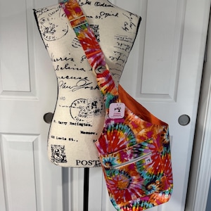 May include: A tie-dye shoulder bag with a zippered front pocket and adjustable strap. The bag features a vibrant pattern of red, orange, yellow, blue, and green swirls. The interior lining is orange.