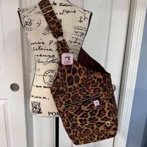 May include: A leopard print crossbody bag with a brown interior and strap. The bag features a zippered pocket, a metal ring, and a small tag that reads "CYNDEE DESIGNS". The bag is displayed on a mannequin.