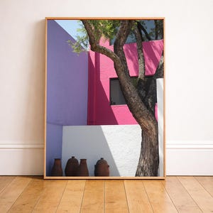 Könnte beinhalten: Gerahmter Druck mit einer lebendigen Architekturszene. Ein Baum mit strukturiertem Stamm und grünen Blättern steht vor einem Gebäude mit Wänden in Rosa-, Lila- und Weißtönen. Braune Töpferwaren stehen vor der weißen Wand.