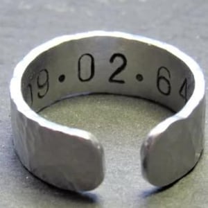 May include: Silver adjustable ring with the engraved date "19.02.64". The ring has a hammered texture and an open design. A piece of jewellery.