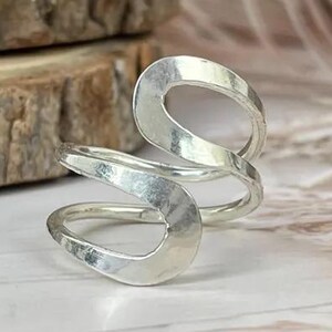 May include: A silver ring with a unique, open design. The ring has two oval shapes connected by a curved band. The metal has a hammered texture. The ring is displayed on a light-coloured wooden surface.