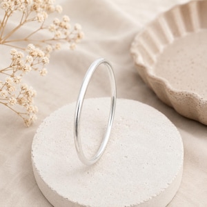 Puede incluir: Un brazalete plateado de diseño liso y redondo. Se exhibe sobre un pequeño pedestal circular de hormigón claro. El fondo incluye una tela beige y un plato decorativo. El brazalete tiene un diseño simple y minimalista.