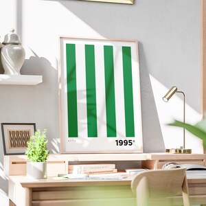 Puede incluir: Un estampado enmarcado con un dise&ntilde;o minimalista con rayas verticales verdes y blancas. El n&uacute;mero "1995" est&aacute; impreso en negro en la parte inferior derecha. El estampado se exhibe en un escritorio de madera con una peque&ntilde;a planta y una l&aacute;mpara de escritorio dorada.