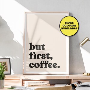 May include: A framed print with the text "but first, coffee." in a bold, black font. The print has a light beige background and is displayed in a light wood frame. A yellow circle with the text "MORE COLOURS AVAILABLE" is in the upper right corner.