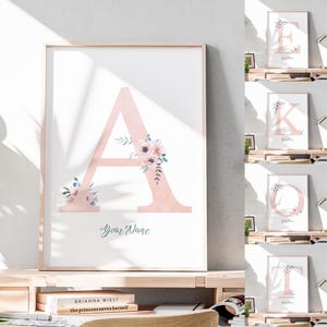 May include: A framed print displaying a large, pale pink letter "A" decorated with floral details. The artwork includes the text "Your Name" in a script font. Other prints with the letters E, K, O, and T are also visible.