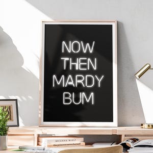 May include: A framed art print with the words "NOW THEN MARDY BUM" in white neon-style lettering against a black background. The print is in a wooden frame and sits on a wooden desk. A small framed picture and a potted plant are also on the desk.