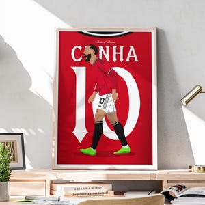 Puede incluir: Una impresión artística enmarcada que presenta una ilustración estilizada de un jugador de fútbol con una camiseta roja con el nombre "CUNHA" y el número "10" en blanco. El jugador está en movimiento, con zapatos verdes y calcetines negros.