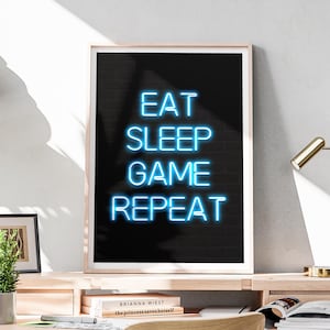 May include: A framed art print featuring the text "EAT SLEEP GAME REPEAT" in bright blue neon-style lettering against a black brick backdrop. The frame is light wood, and the print is displayed on a wooden desk.