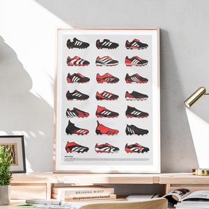 May include: A framed print featuring a collection of soccer cleats in various designs. The cleats are primarily black, red, and white, arranged in a grid pattern against a light gray background. The print is displayed on a wooden surface.
