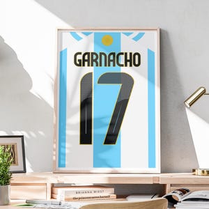 Könnte beinhalten: Gerahmter Druck eines argentinischen Fußballtrikots. Das Trikot ist weiß und hellblau gestreift mit dem Namen "GARNACHO" in Schwarz und der Nummer "17" in Schwarz mit goldener Umrandung. Oben befindet sich ein gelber Kreis.